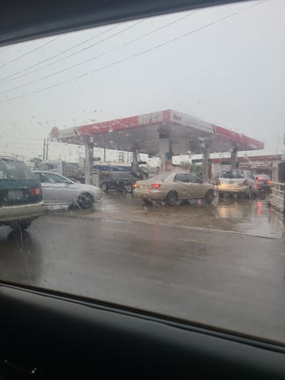 This image depicts a gas station scene in Karu, Nigeria, during what appears to be a rainy day. The perspective is from inside a vehicle, looking out through a wet window, with raindrops visible on the glass. Several cars are lined up at the gas pumps under a large, red and white canopy. The ground is wet and reflective, indicating recent or ongoing rain.
The most prominent object is the gas station structure with its canopy and pumps. Cars are the primary subjects, with at least three visible in the queue. The cars are of various colors and models, but none are actively fueling up at this moment. The overall atmosphere is somewhat dreary due to the weather.
The sky appears overcast, suggesting it's daytime, likely late morning or afternoon, though the exact time is difficult to ascertain due to the weather conditions. The presence of cars waiting at a gas station suggests an activity of refueling or perhaps a queue due to limited services or high demand.
Visible text includes "NIPCO" on a sign and what appears to be "Kano" on another sign. The context of Karu, Nigeria, is provided. The image captures a mundane, everyday scene of a gas station under adverse weather conditions, with the rain adding a layer of atmosphere and altering the visual quality of the scene. The focus is on the vehicles and the infrastructure of the gas station, viewed through the slightly obscured lens of a wet car window.