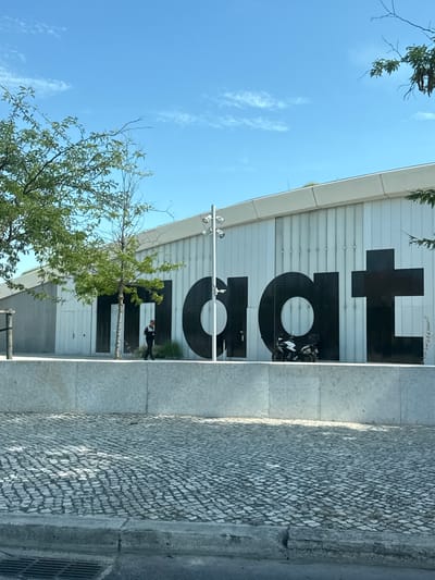 The image captures a frontal view of the MAAT (Museum of Art, Architecture, and Technology) building in Lisbon, Portugal, on a clear day. The museum's facade is characterized by large, black sans-serif letters spelling out "MAAT". The building itself features a white, corrugated metal exterior.
In front of the building, a person in dark clothing is walking towards the right side of the frame. To the right of this person and in front of the "a" and "t" of "MAAT", a white and black motorcycle is parked.
The foreground of the image is dominated by a cobblestone street, with a concrete curb and a metal drain grate visible at the bottom. A low, grey concrete wall runs horizontally across the middle of the frame, separating the street from the museum's entrance area. Trees with green foliage are present on the left and right sides of the image, framing the view of the building. The sky is bright blue with a few wispy clouds.