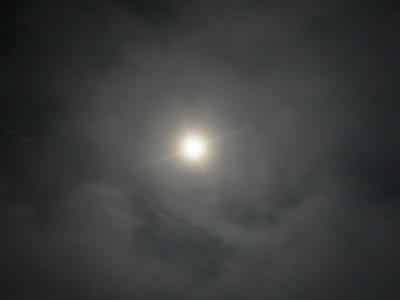 The image depicts a night sky with the moon partially obscured by clouds. A bright, glowing orb, likely the moon, is visible in the upper central portion of the frame, radiating light. Wispy clouds surround the moon, creating a hazy effect. Several thin, dark lines traverse the image diagonally from the lower left to the upper right, suggesting power lines or wires. The overall lighting is dim and atmospheric. The image provides no visual information about the city of Kuru Karama, Nigeria.