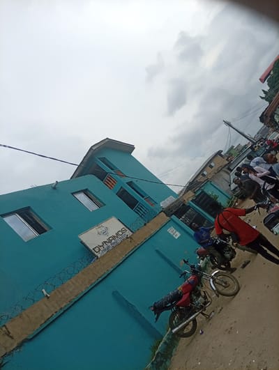 The image depicts an overcast daytime scene on a bustling urban street in Ijanikin, Nigeria. A prominent multi-story building, painted a striking teal or turquoise, occupies the left side of the frame. Affixed to its facade is a sign that clearly reads "DYNAMOS NURSERY PRIMARY AND COLLEGE," with a crest above the text and smaller, partially visible text below that appears to say "COMPUTER COLLEGE." A line of barbed wire runs along a concrete ledge on the lower section of the building, indicating a security measure.
The street in the foreground is unpaved, appearing as dirt or gravel. Several people are present, interacting with one another or with motorcycles. A man in a red shirt stands prominently next to a red motorcycle in the mid-ground, while another man in a blue shirt is visible further back, also near a motorcycle. Other individuals are gathered further down the street. At least three motorcycles, likely commercial "okadas," are visible, either parked or in use, contributing to the busy street ambiance.
The sky is uniformly grey and cloudy, suggesting an overcast day. In the background and to the right, a utility pole with overhead power lines is visible, along with glimpses of other buildings and vehicles, confirming an active urban setting. The overall scene portrays a typical street environment in a Nigerian town or city.