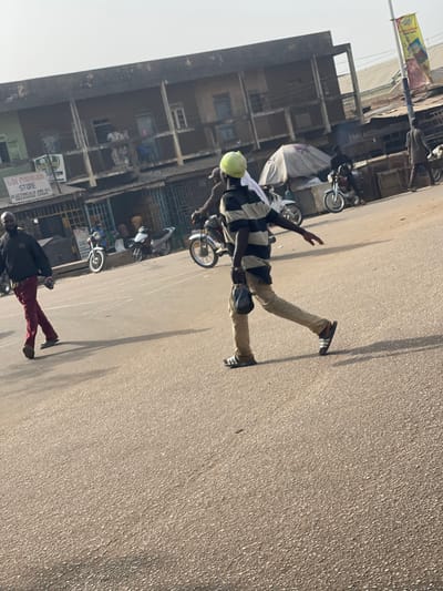 The scene depicts a sunlit street in Chikaji, Nigeria. A male subject is observed in the mid-ground, walking from the right to the left across a paved road. He wears a light-green head covering, a horizontally striped t-shirt (dark and light bands), light-colored trousers, and dark slides with white stripes. He carries a dark, possibly plastic, bag in his left hand, and his right arm is extended laterally. To the left of this individual, another male subject walks from left to right, dressed in a dark long-sleeved shirt and red trousers.
In the background, a two-story building with a weathered facade features multiple windows on its upper floor and several commercial shops at ground level. One visible shop sign reads "GDI PROVISION STORE," with additional text "ELECTRICALS AND ALL RECHARGE CARDS." Several motorcycles are present in the mid-ground and background; some are parked near the shops, and others appear to be in motion or stationary with riders. A light-colored canopy is visible further right in the background, partially obscuring a structure. Another male subject, wearing dark clothing, walks away from the viewer on the far right. A tall, multi-segment pole with a yellow and white banner stands on the right side of the frame. Long, distinct shadows are cast across the asphalt road surface, indicating direct overhead sunlight.