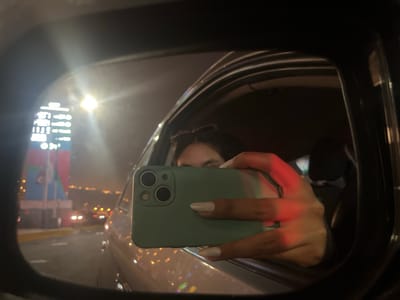 The image depicts a person holding a smartphone up to a car's side mirror, presumably to take a photo or video. The scene reflects in the mirror, showing a blurry view of a brightly lit sign and surrounding lights, typical of an urban nightscape. The location is identified as Lurín, Peru, due to the presence of a distinctive tall sign visible in the background. The individual is visible behind the smartphone, and the interior of the car is partially visible, suggesting they are seated inside. The focus is on the reflection captured in the side mirror, with the smartphone and hand obscuring part of the view. The environment suggests an urban setting at night, with street lights and other vehicles visible in the background.