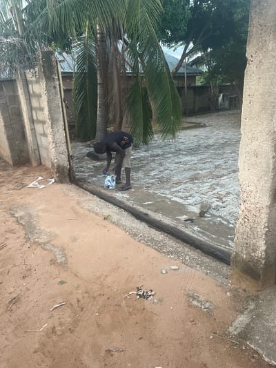 A man in a black t-shirt and patterned shorts is pouring oil from a white container onto a paved area next to a curb. The location appears to be outdoors, with a palm tree and other foliage nearby. There is evidence of water on the paved surface, and the adjacent ground is sandy and uneven. The location might be in Makurdi, Nigeria, but that cannot be forensically confirmed from the image.