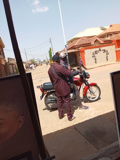 Two men are on a red motorcycle parked on a dirt road. The man on the back wears a red plaid suit and sandals. The driver wears a baseball cap, jacket, gray pants, and blue flip-flops. A white pole stands near the motorcycle. In the background, there are buildings with orange roofs and walls. People walk in the distance. The location is in a city with sandy ground, likely Yelwa, Nigeria.