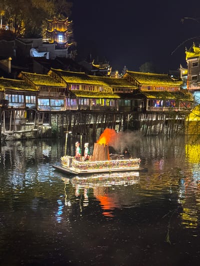 The image depicts a nighttime scene on a river in Fenghuang, China. A rectangular, flat-bottomed boat, adorned with colorful flowers and perimeter lighting, floats on the dark, reflective water. Onboard, two individuals stand prominently towards the front-left, wearing traditional, multi-colored garments and headwear. A tall, conical wooden structure on the boat emits bright orange flames and dark smoke. A third individual in dark attire is partially visible on the right side of the boat. The boat's lights and the fire's glow are reflected on the water's surface.

Along the riverbank in the background, a series of multi-story traditional Chinese stilt houses (diaojiaolou) are brightly illuminated with warm yellow lights along their eaves and windows. These wooden structures are supported by numerous stilts extending into the river. Higher on the hill behind the stilt houses, a prominent, multi-tiered pagoda-style building with illuminated windows and traditional curved roofs is visible against the dark sky. Scattered illuminated trees and additional buildings can be seen on the hillside. A slender, dark branch hangs into the water from the right side of the frame.