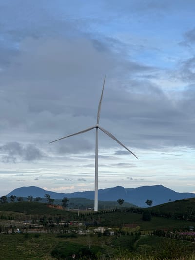 The image shows a wind turbine in Phát Chi, Vietnam. The turbine is white with three blades and stands tall against a backdrop of cloudy skies. In the distance, mountain ranges are visible, and the land around the turbine appears to be lush with green vegetation and tea plantations. The photograph is taken from a slightly low angle, emphasizing the height of the turbine.