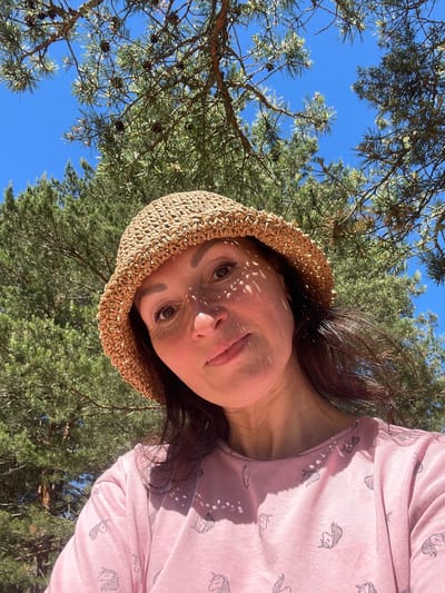 A woman with dark hair and fair skin wears a woven straw hat and a pink t-shirt with a unicorn print. She is outdoors, under a canopy of pine trees with visible pinecones, and against a bright blue sky. Sunlight filters through the needles, casting dappled shadows on her face and t-shirt. The scene appears to be a snapshot taken in Zelenaya Roshcha, Russia, on a sunny day.