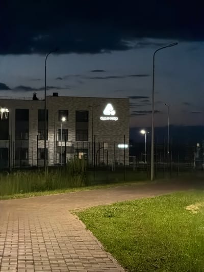 An exterior, nighttime, eye-level, medium shot shows a modern, brick building with a large, illuminated logo in white on its facade. The logo is a stylized triangle with a smaller triangle inside it, and the Cyrillic text "адымнар" is positioned below it. The building appears to be a school or community center, with multiple windows visible, some illuminated from within. The foreground features a paved walkway made of rectangular bricks, bordered by a strip of green grass and then taller green vegetation. In the background, tall metal structures, possibly sports equipment, are visible behind a fence. Several streetlights are present, casting light on the scene. The sky is dark with ominous, heavy clouds. The setting is identified as being in the city of Naberezhnye Chelny, Russia.