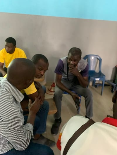 An indoor scene in Fagge A, Nigeria, depicts five adult males engaged in a discussion or meeting. The room features a two-toned wall with a light blue upper section and a light grey lower section, above a light-colored floor. In the foreground, a bald man in a white and black striped long-sleeved shirt faces away from the camera, gesturing with his right hand towards a man in a yellow t-shirt and blue jeans, who is looking down. To the right, another man, wearing a grey and purple short-sleeved polo shirt and dark grey trousers, is seated on a blue plastic chair, holding a dark tablet or phone in his left hand, with his right hand near his chin, observing the interaction. In the background, a third man in a yellow t-shirt is partially visible, seated. A second blue plastic chair is empty in the background, and a small red and white object rests on the floor. A person in a white garment with a brown stripe is partially visible in the extreme foreground, right.