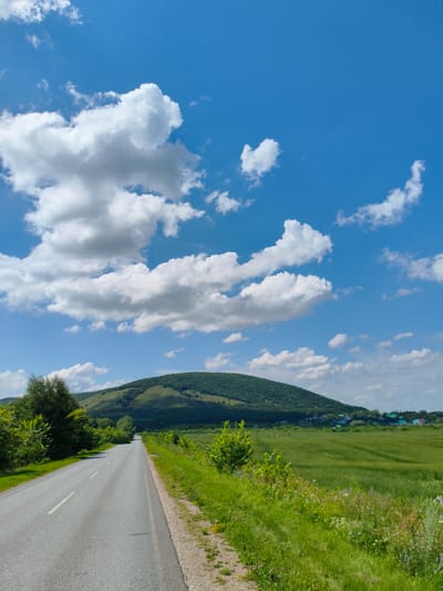 An asphalt road stretches into the distance under a bright blue sky filled with fluffy white clouds. The road is lined with lush green grass and trees. To the right of the road is a large field of green crops. In the background, a rolling green hill is visible, and a small village with colorful roofs can be seen nestled at its base. The image appears to be taken in Bel'skoe, Russia, on a clear, sunny day. There are no people or animals in the image, and no specific actions are taking place. The environment is rural and agricultural.