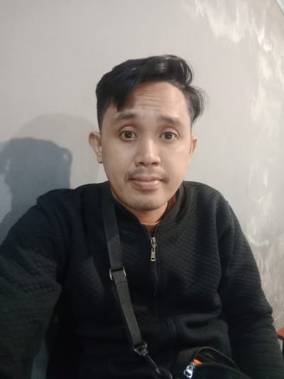 A medium-shot portrait depicts an adult male of apparent Southeast Asian descent, positioned centrally and looking directly forward. He has short, dark, styled hair, dark eyes, a thin mustache, and light stubble on his chin and upper lip. The subject wears a black, quilted, zip-up jacket with a silver-toned zipper, partially unzipped to reveal a thin, light-colored necklace or chain. A black fabric strap, consistent with a shoulder bag or camera, diagonally crosses his left shoulder and chest. A section of a dark bag with an orange zipper pull is visible in the lower right foreground, connected to the strap. The background consists of a plain, light gray or off-white wall exhibiting minor textural inconsistencies and a pronounced dark shadow cast to the left of the subject. The lighting appears to be artificial. The specific location within the city of Semarang, Indonesia, cannot be determined from the visual information provided in the image.