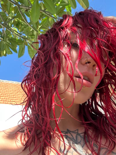 A low-angle shot depicts a person with red hair, possibly in Dalung, Indonesia. The perspective is from below, looking up at the individual. Visible are the person's face, including lips, nose ring, and one eye, obscured by strands of red hair. Part of a tattoo is visible on their shoulder. The background includes green leaves from a tree, a patch of blue sky, and a glimpse of a building with brickwork.