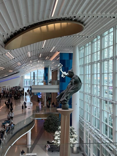 The image depicts the interior of a modern airport terminal. A large, abstract sculpture of a robed figure is prominently displayed, leaning forward and holding what appears to be a glowing object. The terminal features a sloped, textured wall with integrated lighting on the left side, and a vast glass ceiling that allows natural light to filter in, revealing the building's structural framework above.

People are visible throughout the scene, some seated on benches or luggage, others walking or standing in groups. They appear to be travelers, as suggested by the presence of luggage and the general ambiance of an airport. The overall atmosphere suggests a busy travel hub, likely during daylight hours, given the ample natural light.

The floor is highly reflective, mirroring the ceiling and people, adding to the sense of spaciousness and modern design. In the background, there are signs and indications of different sections of the airport, including escalators and seating areas. The blue tiled wall behind the sculpture adds a pop of color to the otherwise neutral palette of the interior.

While no specific time of day is explicitly stated, the bright, diffused light suggests it is daytime. The weather outside is not visible. The location is identified as Charlotte, USA. There is text visible on a blue sign that reads "ALL GATES" and another sign with an arrow and "EXIT 1".
