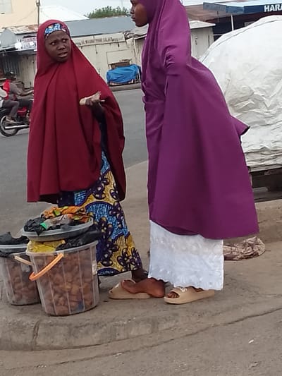 Two women are standing on a sidewalk in Pantame, Nigeria. The woman on the left is wearing a dark red hijab and a colorful patterned skirt. She is holding a small object in her hand and looking to her left with a surprised expression. To her right, a woman is wearing a purple hijab and a white skirt. In front of the women are two clear plastic buckets filled with food items. The sidewalk is made of concrete, and there is a street behind them with a motorcycle and buildings in the background. The sky is overcast.