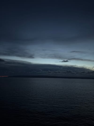 The image displays a twilight seascape observed from an elevated vantage point in Punta de Maisi, Cuba. The upper two-thirds of the frame are dominated by a cloudy sky, transitioning from dark blue-gray at the zenith to a lighter, pale blue-gray horizontal band just above the distant horizon. Various darker, layered cloud formations are visible, with some smaller, discrete clouds against the lighter sky band. A distinct, dark, horizontal line forms the horizon, separating the sky from the water. A low, dark, silhouetted landmass, consistent with a distant coastline or island, is visible along this horizon. On the far left, a faint, localized orange-red light source is present just above the landmass and below the clouds. The lower third of the image is occupied by a vast expanse of dark blue-gray water, exhibiting minor surface ripples, indicative of a calm sea or ocean during low light conditions. No individuals, vessels, or man-made objects are visible within the frame. The scene is a natural coastal environment under overcast, low-light conditions.