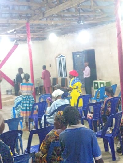 Inside a brightly lit church with exposed ceiling beams, people sit on blue plastic chairs. Several individuals stand near the front, including a man in a pink shirt and a woman in a yellow dress with a red headscarf. A window with bars is visible behind them. Red fabric decorates the support beams. The room appears modest and crowded with attendees.