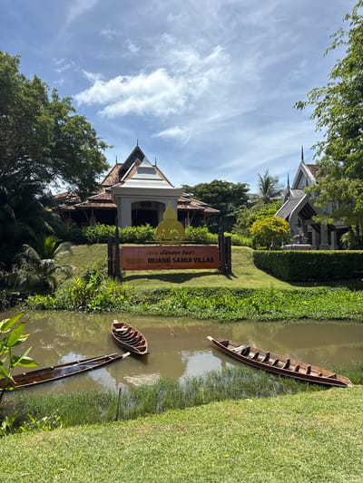 In the foreground, a narrow, muddy canal is visible, with lush green grass lining its banks. Three wooden rowboats are partially submerged in the water. The middle boat is longer and appears to be leaning to one side. The boat on the right is shorter and also sits low in the water. In the background, a sign reads "ROYAL MUANG SAMUI VILLAS" in English. Behind the sign, a building with a traditional Thai architectural style features a pitched, tiled roof. The building is surrounded by greenery, including trees and manicured bushes. A golden Buddha-like statue is visible above the sign. The sky is blue with scattered white clouds.