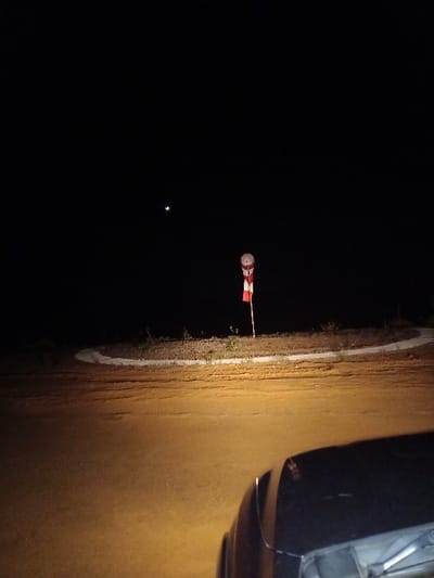 The scene depicts a nocturnal outdoor environment in Santa Juana, Chile. The foreground is illuminated by a warm, yellowish light source, originating from a dark-colored vehicle whose hood and front fender are partially visible in the lower right. This light falls upon an unpaved ground surface, likely a dirt or gravel road. In the midground, a raised, curving median or island, covered with sparse vegetation, is present. A vertical pole stands centrally on this median, characterized by distinct red and white horizontal stripes. Mounted at the top of the pole is a white windsock, featuring a prominent red band near its opening, affixed to a circular frame. The background consists of a dark night sky, with a visible crescent moon positioned in the upper left quadrant. No individuals, explicit actions, or direct interactions are discernible within the frame.