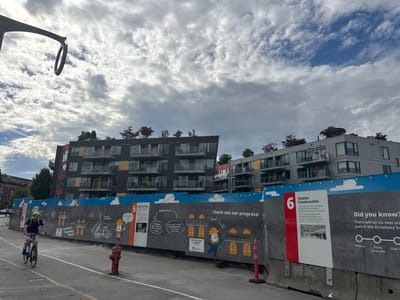 The image shows a man cycling past a construction site in Vancouver, Canada. The construction site has a large mural on a temporary barrier that depicts workers and information about the Broadway Subway project. The mural has several sections with text and illustrations, including a section titled "Station Construction" and another titled "Check out our progress!". The man on the bicycle is wearing a yellow helmet and a backpack. In the background, there are modern apartment buildings. The sky is cloudy.