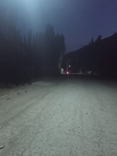 This is a nighttime, low-light, horizontal, eye-level shot of a dirt road in the city of Sayod, Tajikistan. The road is dusty and uneven, with scattered rocks. To the left of the road, there is a dark, forested area, with tall, slender trees silhouetted against a hazy, blue sky. The sky is overcast and there is a bright light source in the upper left quadrant of the image, possibly a streetlight or the moon. In the distance, the road curves to the right, leading towards a dimly lit area where a few buildings and a vehicle are visible. The overall mood is dark and somber, with a sense of isolation.