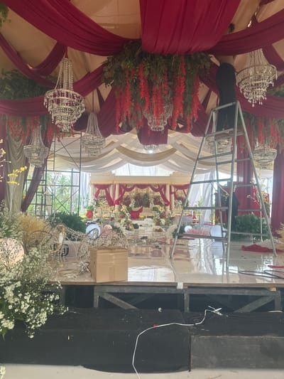 The image captures an indoor event setup, likely a wedding reception or banquet hall, in Aluebilirayeuk, Indonesia. The scene is adorned with elaborate decorations, featuring draped burgundy and cream fabric along the ceiling, interspersed with lush green foliage and cascading red floral arrangements. Several ornate crystal chandeliers are suspended from the ceiling, adding a touch of grandeur. In the foreground, a ladder is positioned, with a person on it partially obscured by the structure, appearing to be engaged in the setup or decoration process. The floor is a polished, reflective surface, showing some scattered debris and wires, indicating ongoing preparations. In the background, a stage area is visible, decorated with more floral arrangements and a seating area. The overall ambiance suggests a formal and festive event in progress or nearing completion, set within a tropical or sub-tropical environment, hinted at by the greenery visible through the open sides of the structure.
