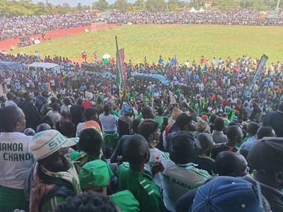 The image captures a large, bustling political rally or public gathering within an outdoor stadium in Siaya, Kenya, during the daytime. The stadium features a red running track encircling a green grass field, both densely packed with attendees.
In the foreground, the backs of numerous individuals are visible, many wearing green shirts, caps, or scarves, suggesting a shared affiliation. One person wears a white t-shirt with partially visible text "HANDA CHANDO," and another dons a white cap with "SIEL." Another individual's t-shirt reads "FOR MARIA."
The midground reveals a crowded section on the stadium track, where a makeshift stage or platform is set up with large speakers. Several individuals are standing on this platform, seemingly addressing the crowd. A white van or bus is parked nearby. Flags and banners, predominantly green with some red, are held aloft, with partial text like "OPOYO" visible on one green banner.
Beyond this central activity, the stadium stands are completely filled with a vast multitude of people, extending into the background. The atmosphere appears lively and engaged, with many onlookers facing the stage area. The sky is bright, indicating sunny weather. The overall scene conveys a large-scale public event with significant attendance.
