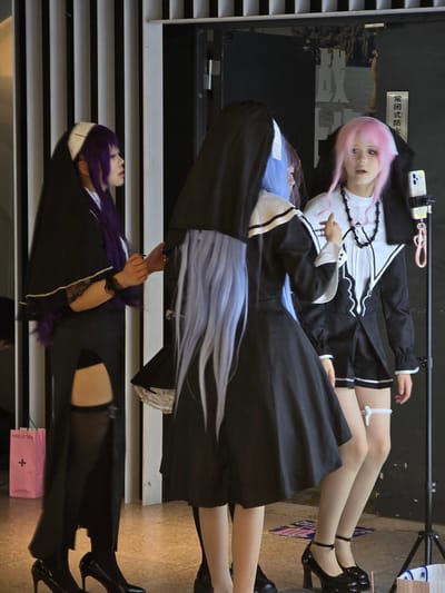 In Shangcheng District, China, three individuals are posing for a photograph. The person on the left has purple hair and is wearing a black nun-like costume with a white wimple. The person in the middle has light blue hair and is wearing a similar black nun-like costume. The person on the right has pink hair and is wearing a black and white uniform with a necklace. They are standing in front of a black door with white vertical blinds on the left side. A pink shopping bag with "MOLLY TEA" written on it is visible on the floor to the left. A phone mounted on a tripod is positioned to the right of the person with pink hair.