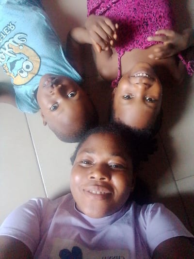 The media file is a close-up, overhead shot of three people lying down, seemingly posing for a selfie. Two children are at the top of the frame, positioned with their heads touching. The child on the left is wearing a light blue t-shirt with a graphic on it. The child on the right is wearing a vibrant pink, textured top. The third person, an adult, is positioned at the bottom of the frame, with their head and upper torso visible. They are wearing a light purple t-shirt. All three subjects are smiling and looking towards the camera, suggesting a positive and joyful mood. The background appears to be a tiled floor, a common setting in many homes. The lighting suggests it is daytime. The image was taken in Yenegoa, Nigeria. There is some visible text on the blue t-shirt but it is partially obscured and difficult to read.