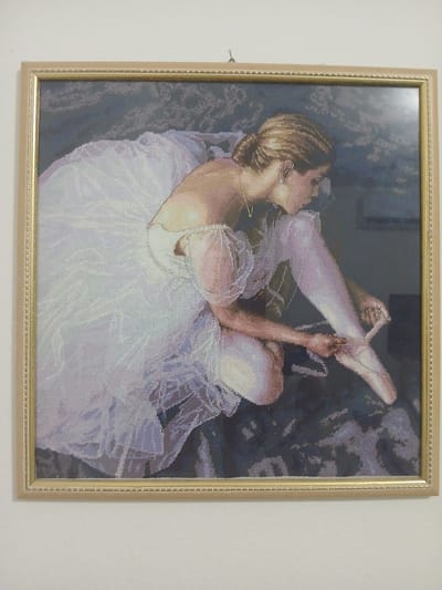 This framed cross-stitch depicts a young woman, likely a ballet dancer, in a white tutu and pointe shoes. She is shown from the side, seated, and is in the process of tying the ribbons of her pointe shoe. Her head is tilted back, and her eyes are closed, suggesting concentration or a moment of repose. The background is dark and abstract, with hints of deep blue and purple, creating a dramatic contrast with the light colors of the dancer's attire. The cross-stitch has a soft, pixelated appearance typical of the medium. The frame is a simple, light-colored wood with a beaded edge. There is no visible text in the image. The scene evokes a sense of quiet dedication and the beauty of ballet.