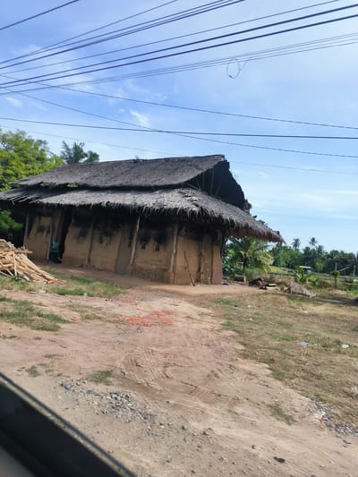 The image depicts a rustic, thatch-roofed structure with mud brick walls, possibly a dwelling or workshop, situated in a rural area. A person in a blue shirt stands within an open doorway of the structure, partially visible. Piles of wooden poles and scattered bricks are present in the foreground, suggesting ongoing construction or material storage. The surrounding environment is characterized by dry, unpaved ground, sparse vegetation, and a backdrop of lush green trees and palm trees, indicative of a tropical climate. The scene is further characterized by a network of overhead power lines against a clear blue sky with scattered clouds. The overall impression is one of a simple, traditional settlement in an undeveloped or developing region. The specific location is Lancang Barat, Indonesia.