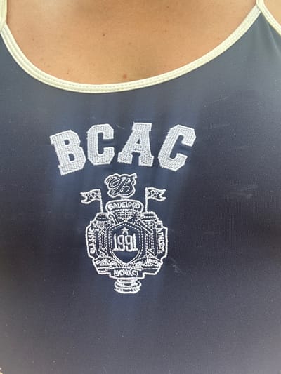 The media file shows a close-up of a dark blue garment, possibly a swimsuit or athletic wear, with white embroidered text and a crest. The garment has a light-colored trim along the edge. The embroidery on the left reads "BCAC" in large, block letters. To the right of the text is a circular crest with intricate details. Inside the crest, the number "1991" is prominently displayed. Surrounding the number and forming the crest are various other embroidered elements, including what appears to be text and decorative patterns. The overall impression is of branded athletic apparel, likely associated with an organization or event from 1991. The background is dark and out of focus, drawing attention to the embroidered design on the garment. The image is well-lit, highlighting the texture of the embroidery and the fabric. No specific activity or event is depicted, and no location cues are visible beyond the garment itself. The context provided of "Aiyrtau, Kazakhstan" does not correlate with any visual information within the image.