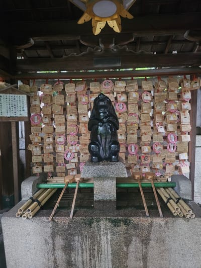 This image captures a purification area at Okazaki Shrine in Kyoto, Japan. Centrally positioned is a large, dark, glossy, stylized statue of an upright rabbit resting on a grey stone pedestal. Behind the statue, a wooden lattice wall is densely covered with numerous rectangular wooden votive tablets (ema), many featuring illustrations of two white rabbits enclosed within a pink circular rope design. A wooden sign on the left, written in Japanese, confirms the location as Okazaki Shrine and provides instructions for the ritual, indicating a wish-making process involving pouring water on the rabbit.

In the foreground, a rectangular stone purification basin shows signs of water streaks and wear. Two wooden ladles (hishaku) with long handles are placed across a green pipe or channel extending into the basin. On either side of the basin, bundles of natural bamboo poles are stacked horizontally. Overhead, dark wooden architectural elements with exposed beams and carved details are visible. A golden, ornate ceiling fixture with a central circular light source is positioned at the top center of the frame. No individuals are present in the scene.