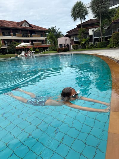 A young person with goggles and patterned swim trunks swims freestyle in a turquoise swimming pool. They are submerged to their chest, with arms extended forward. The pool has a tiled bottom with a visible grid pattern. To the left, a resort-style building with red tile roofs and balconies is visible, surrounded by palm trees and other tropical foliage. Several lounge chairs and a white umbrella are set up on the poolside. In the background, other people can be seen relaxing by the pool. The sky is overcast.