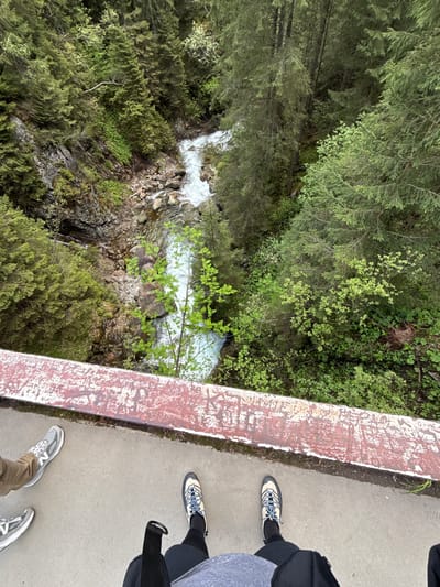 The image shows a view from a bridge overlooking a stream in a heavily forested area. Two individuals are visible on the bridge, with their feet and lower legs appearing in the frame. The bridge has a weathered, reddish-brown painted barrier. The stream below is rocky and the water is turbulent. The surrounding vegetation is lush and green. There are no recognizable landmarks or signs indicating this is Brzegi, Poland.