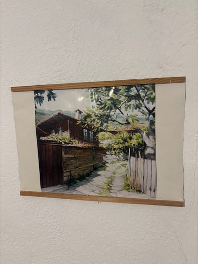 A watercolor painting depicts a quaint street scene in Shipkovo, Bulgaria. The artwork captures a rustic wooden house with a shingled roof, set against a backdrop of rolling hills. In front of the house, a cobblestone pathway leads past a low wooden fence, bordered by lush green foliage. The painting evokes a sense of tranquility and traditional Bulgarian architecture. The painting is framed with a simple wooden border. The wall on which the painting is hung has a textured, white finish with visible cracks.