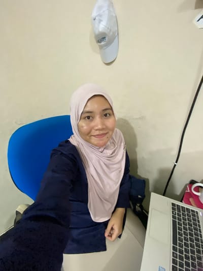 In a room, a young woman wearing a light pink hijab and a dark blue shirt is seated in a bright blue chair, facing the camera. She has fair skin with some blemishes on her cheek and a mole on her chin. She is smiling slightly. To her right, a laptop is visible, open and positioned on a surface. Behind her, a white cap with a logo is affixed to a light-colored wall. The overall impression is that of a casual indoor setting, possibly a home office or study area, in Banjarbaru, Indonesia.