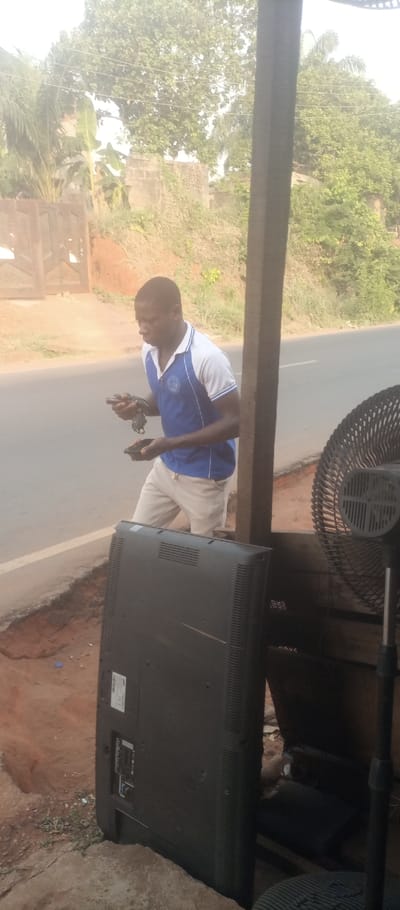 The image depicts an outdoor scene in Ogidi, Nigeria, capturing a male individual engaged in an activity alongside a road.
In the mid-ground, a dark-skinned male subject is standing, facing slightly to his left. He wears a white polo shirt with blue sleeves and collar, and light-colored trousers. His hands hold multiple small, dark objects, and his gaze is directed downwards towards them, indicating focused attention or manipulation.
To the immediate foreground and right, a large, dark-colored flat-screen television is positioned vertically on its back, displaying its rear panel with visible ports and ventilation grates. Partially obscured to the right of the television is a black standing fan. A thick, dark, vertical post stands in the mid-right, partially framing the scene.
Behind the male subject, a paved road with a visible white edge line extends across the frame. Further back, the terrain slopes upwards, covered with dense green vegetation, including trees and bushes, some appearing to be palm or banana plants. Sections of unfinished concrete block walls and a wooden gate are discernible through the foliage in the background. The immediate ground in the foreground consists of reddish-brown soil with some loose debris. Overhead, thin wires are visible stretched between trees.