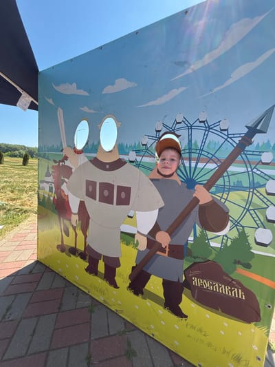A young boy with brown hair and a brown baseball cap is posing for a photo in a cutout board at an outdoor amusement park or fairground. The board depicts two cartoonish figures, one on horseback and another standing warrior, with cutouts for faces. The boy's face is visible through the cutout in the standing warrior's figure. He is holding a long spear. Behind the figures, the background features a Ferris wheel and stylized trees, under a bright blue sky with white clouds. The foreground shows patterned paving stones, with a shadow cast by the cutout board. The scene appears to be located in or near the city of Zavolzhye, Russia, given the presence of a stone with Cyrillic inscription that translates to "Zavolzhye".