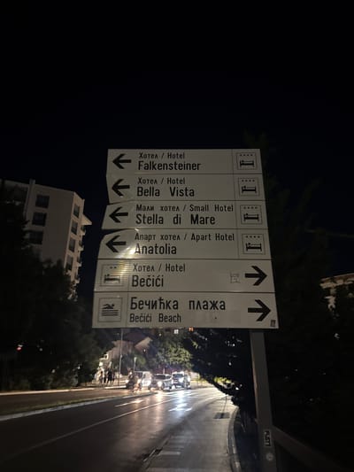 In Rafailovići, Montenegro, at night, a directional signpost stands by a road. The signpost, made of white panels with black text and symbols, displays directions to various hotels and a beach. The hotels listed are Falkensteiner, Bella Vista, Stella di Mare, and Anatolia, each indicated with a left arrow and a hotel icon with four stars. Below these, a sign for Bečići Beach shows a swimming icon and a right arrow, with the text in both Cyrillic and Latin scripts. In the background, a modern, multi-story building is visible, along with trees lining the road. Several cars are driving on the wet road, their headlights illuminating the scene, suggesting recent rain. A few pedestrians are walking on the sidewalk. The sky is dark, indicating nighttime.