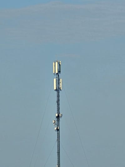 A tall, slender cell tower stands against a clear, pale blue sky. The tower is constructed of dark metal lattice work and features multiple rectangular antennas mounted at various heights. These antennas are primarily white with darker accents. Thin guy wires extend from the tower to the ground, supporting its structure. The background is a uniform sky with subtle, wispy clouds visible towards the top of the frame. There are no people or other objects visible in the frame. The image is a medium shot of the tower, centered vertically and slightly to the right horizontally.