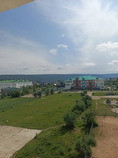 The image depicts a bright, sunny day in Vitim, Russia. In the foreground, a patch of concrete is visible, bordered by a grassy field. A fence runs along the edge of the field, separating it from a sandy area with a volleyball net and playground equipment. Beyond the field, several buildings are visible, including a long, white structure with a green roof and a larger, multi-story building with a distinctive teal roof. The surrounding landscape is characterized by lush greenery and a distant, tree-covered hill. The sky is a clear blue with scattered white clouds. No people are visible in the image.
