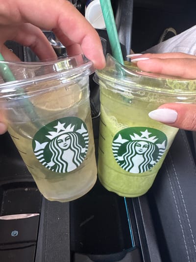 In Arroyomolinos, Spain, two hands hold Starbucks cups, one containing a pale yellow beverage with ice and a lime slice, the other a vibrant green, frothy drink. Both cups display the iconic Starbucks siren logo and are fitted with clear plastic lids and green straws. The image is taken from a close-up perspective, focusing on the drinks and the hands, suggesting the context of a car interior, with a glimpse of black car upholstery and a central console that includes a "P" symbol for parking.