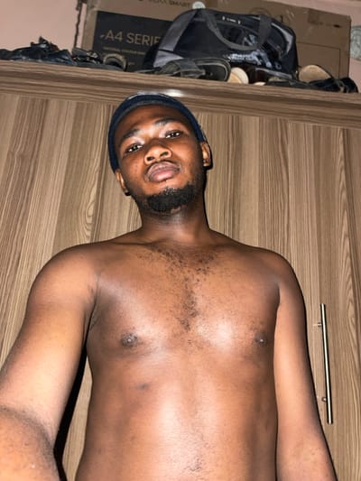 A male subject, appearing to be in his 20s to early 30s with dark skin and a beard, is depicted shirtless, wearing a dark blue or black beanie. He is looking directly at the camera with a neutral expression. His chest displays moderate body hair. The background consists of a wooden cabinet or wardrobe with vertical grain, featuring a metallic pull handle on the right side. Above the subject's head, on top of the cabinet, a cardboard box labeled "A4 SERIES" and "VIDIA SMART" is visible, along with a dark duffel bag and several pairs of footwear. The scene is indoors. This image was taken in Muchiya, Nigeria.