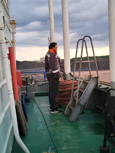 A man stands on a wet, green deck, looking out towards a cloudy sky and a landscape of industrial piles. He is wearing a dark jacket with orange and silver accents, dark pants, and black and white athletic shoes. To his left, a large white pipe runs vertically along the side of a structure. To his right, a metal ladder leads upwards, and a coil of thick, brown rope is visible. In the background, dark industrial mounds are seen against a pale sky. The scene appears to be on a vessel or dockside.