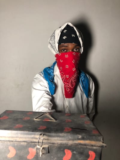A person wearing multiple bandanas, including white, black, red, and blue, stands near a vintage metal box. The person's eyes are visible through the red bandana, which covers the lower half of their face. The white bandana forms part of a hood over their head, revealing a black bandana underneath. The box has a rusted appearance, decorated with red paisley-like designs. A white shirt is partially visible around the person's shoulders. The background is a plain, gray wall.