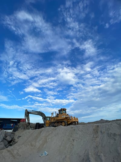 The image depicts a construction or excavation site in Ilado, Nigeria. Dominating the foreground is a large earth mound. A Volvo excavator and a bulldozer are positioned on top of the mound. In the background, there is a dark, container-like structure. The sky is blue with scattered white clouds.
