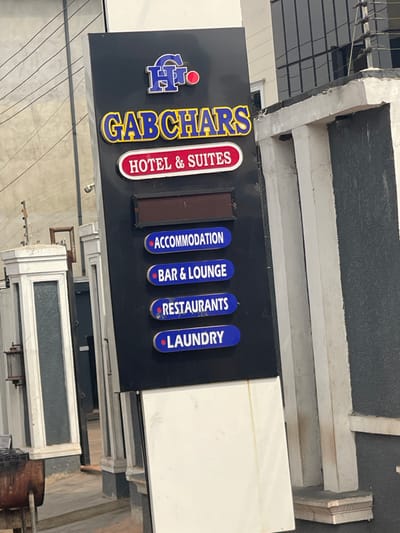A vertical black sign for "GABCHARS HOTEL & SUITES" is prominently positioned in Neni, Nigeria. The sign features a "GH" logo with a red circle at the top, followed by "GABCHARS" in yellow capital letters and "HOTEL & SUITES" within a red-bordered white oval. Below this, an unlit, dark rectangular display panel is present, followed by four blue oval panels listing services: "ACCOMMODATION," "BAR & LOUNGE," "RESTAURANTS," and "LAUNDRY." To the immediate left, a decorative white and grey pillar is visible, with a portion of a metal grill or barbecue apparatus at ground level. Overhead utility lines are visible in the upper left background. To the right, a grey textured wall or pillar abuts the sign, and the dark-tinted windows of a multi-story building are visible above the wall in the background. The ground is a light-colored, paved surface. No individuals are present in the scene.