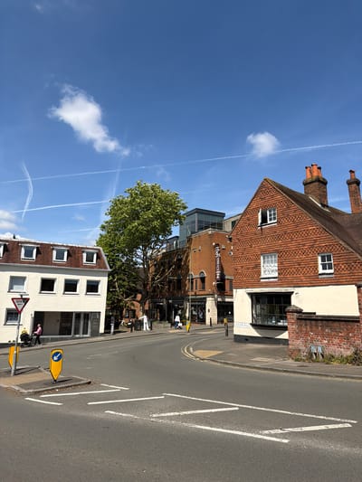 The image depicts a street scene in Guildford, United Kingdom. The view is upwards toward buildings on the side of the road.
Buildings:
- A red brick building with a steeply pitched tiled roof and chimneys, including a white sign that says "SOUTH HILL".
- A modern brick and glass building housing "Thai Terrace" restaurant.
- A white building with multiple rectangular windows and a brown tile roof.
Street:
- An asphalt road marked with white directional arrows and yellow road markings, including a traffic sign that directs traffic to the left.
Other:
- A large green tree partially obscures the view of some buildings.
- A blue sky is visible with several contrails.
- A stroller is on the sidewalk.