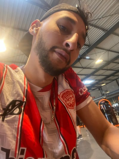 A mid-shot selfie depicts a male individual within an indoor fitness center, specifically identified as a Basic-Fit gym located at 10 Avenue du Général de Gaulle in Hœnheim, France. The subject, a male with a beard and dark hair, wears a black baseball cap reversed and a white wireless earbud in his right ear. He is clad in a red and white striped sports jersey, worn over a white t-shirt, featuring a black "AB" logo on the left chest and a circular emblem on the right sleeve with Arabic text and a mosque motif, along with "COD M". A silver chain necklace is visible around his neck. The background reveals a high industrial ceiling with exposed metallic beams, ductwork, and various lighting fixtures, including both recessed and pendant lights. Exercise equipment with prominent orange accents, including a piece of "MATRIX" branded machinery, is visible to the right of the frame. The floor is a light, uniform color. Faint shapes of other individuals and equipment are discernible in the deeper background.