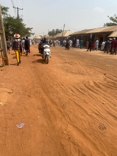 An unpaved, reddish-brown dirt street in Minna, Nigeria, is depicted under bright, hazy daylight. The ground is marked with numerous tire tracks and footprints. On the left, a utility pole with power lines stands beside sparse green trees. Along the right side of the street, a row of single-story buildings with corrugated metal roofs and plastered walls extends into the background. Approximately 30-50 individuals are present on the street. In the foreground on the left, a male subject walks away from the camera, wearing a straw hat, a multi-patterned long-sleeved shirt, and bright yellow trousers. In the center-left, a male subject operates a white scooter-style motorcycle, riding towards the viewer. Another motorcycle with a rider is visible further back on the right. Individuals are seen walking, standing, and riding motorcycles, indicating general street activity or a gathering. Sunlight casts distinct shadows.